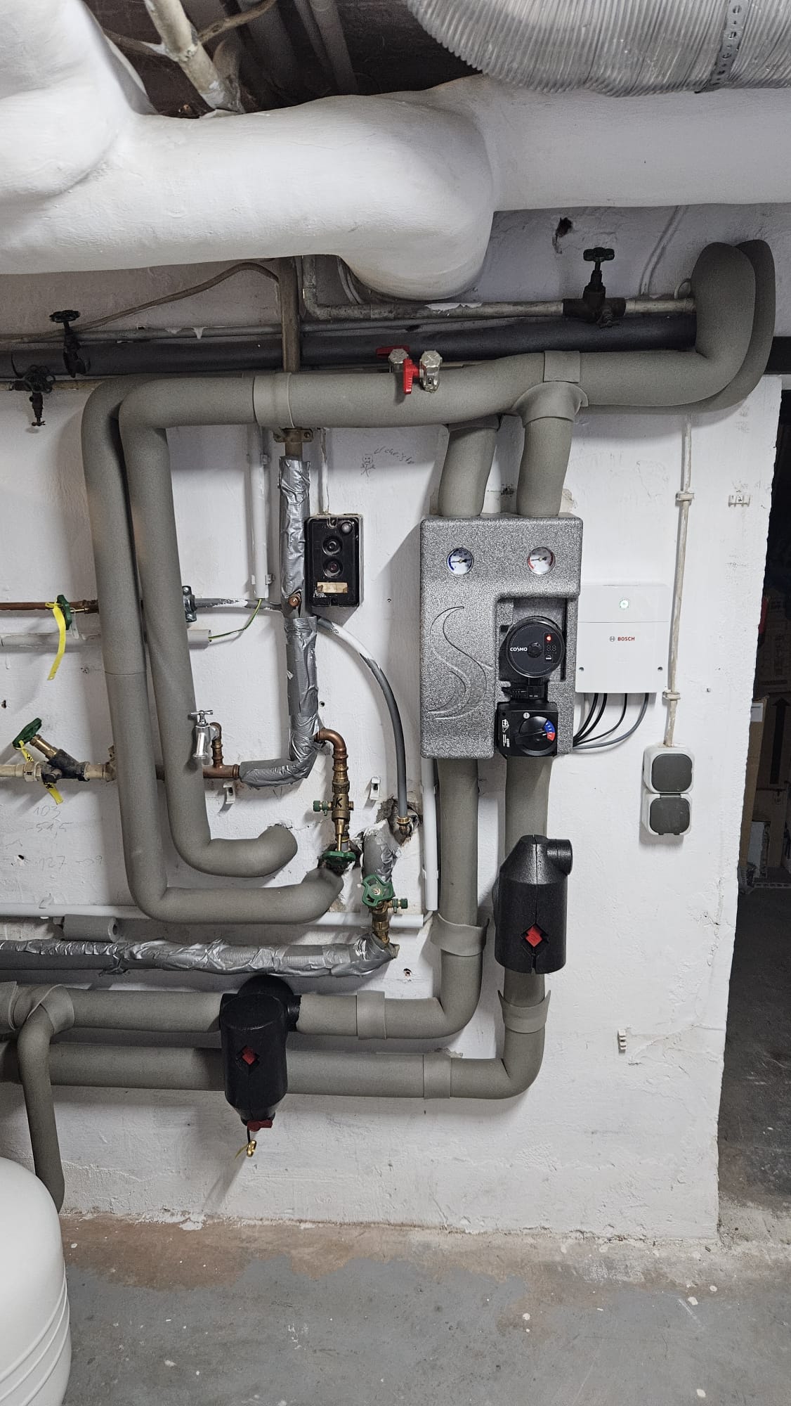 Wand mit komplexer Heizungsrohrinstallation, Isolierungen, Armaturen und einer Pumpengruppe – Teil einer Heizungsanlage.